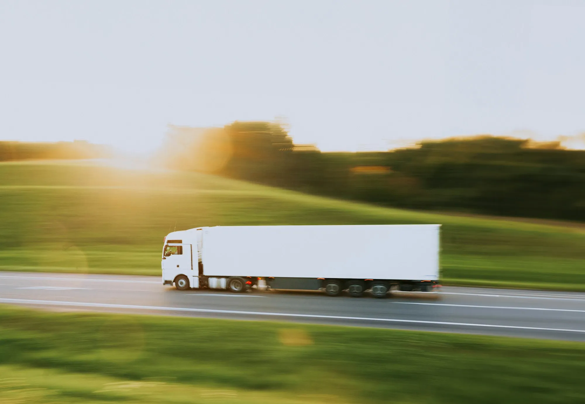 A white semi truck in motion on a country road, golden sunlight flaring across the hills